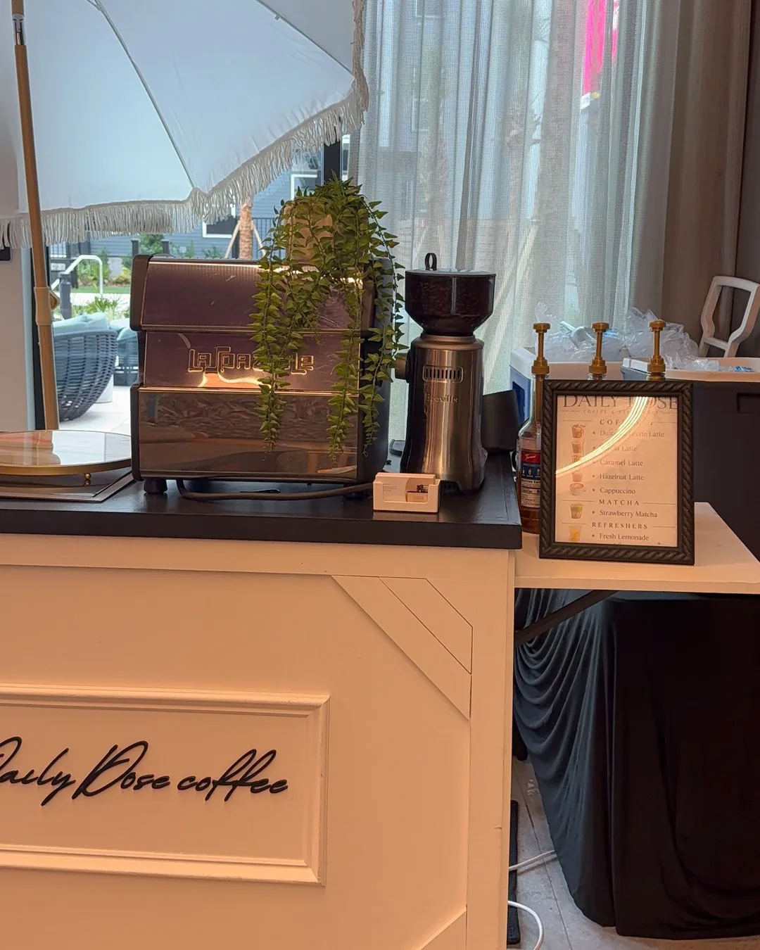 The Daily Dose cart with its cursive sign, a La Spaziale espresso machine and a grinder on the black top, beside a framed menu card listing lattes, strawberry matcha and fresh lemonade