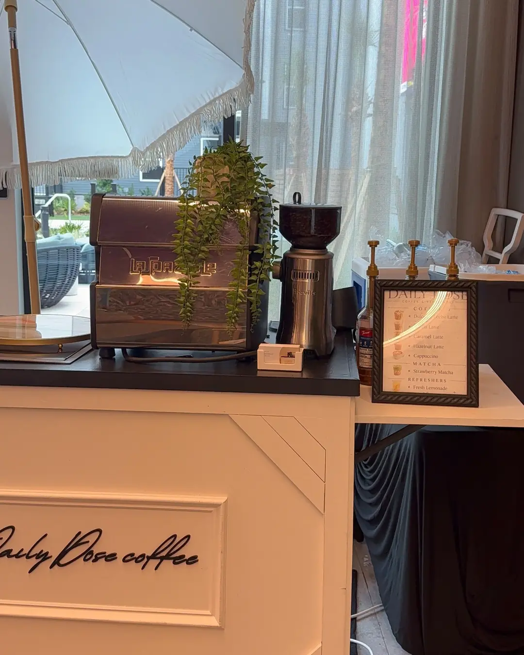 The espresso machine and grinder on the black cart top beside a framed Daily Dose menu card listing lattes, matcha and lemonade