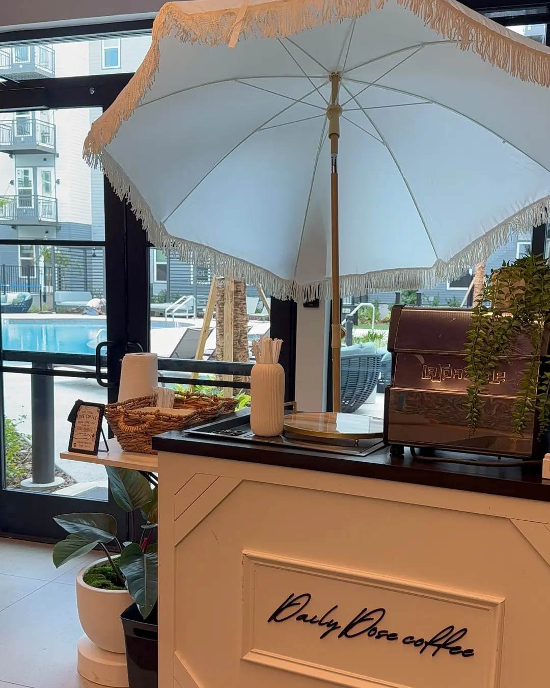 The white Daily Dose cart with its cursive logo, a fringed cream umbrella overhead, and the venue pool through the glass behind it
