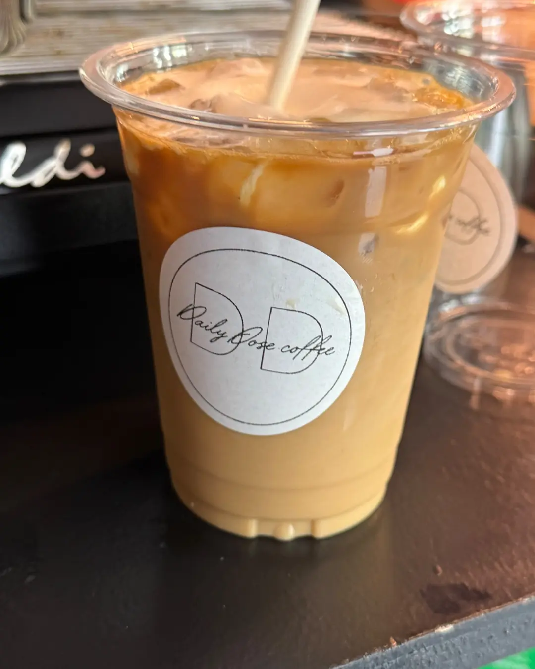 A close-up of an iced latte in a clear cup with the Daily Dose sticker, resting on the black cart top